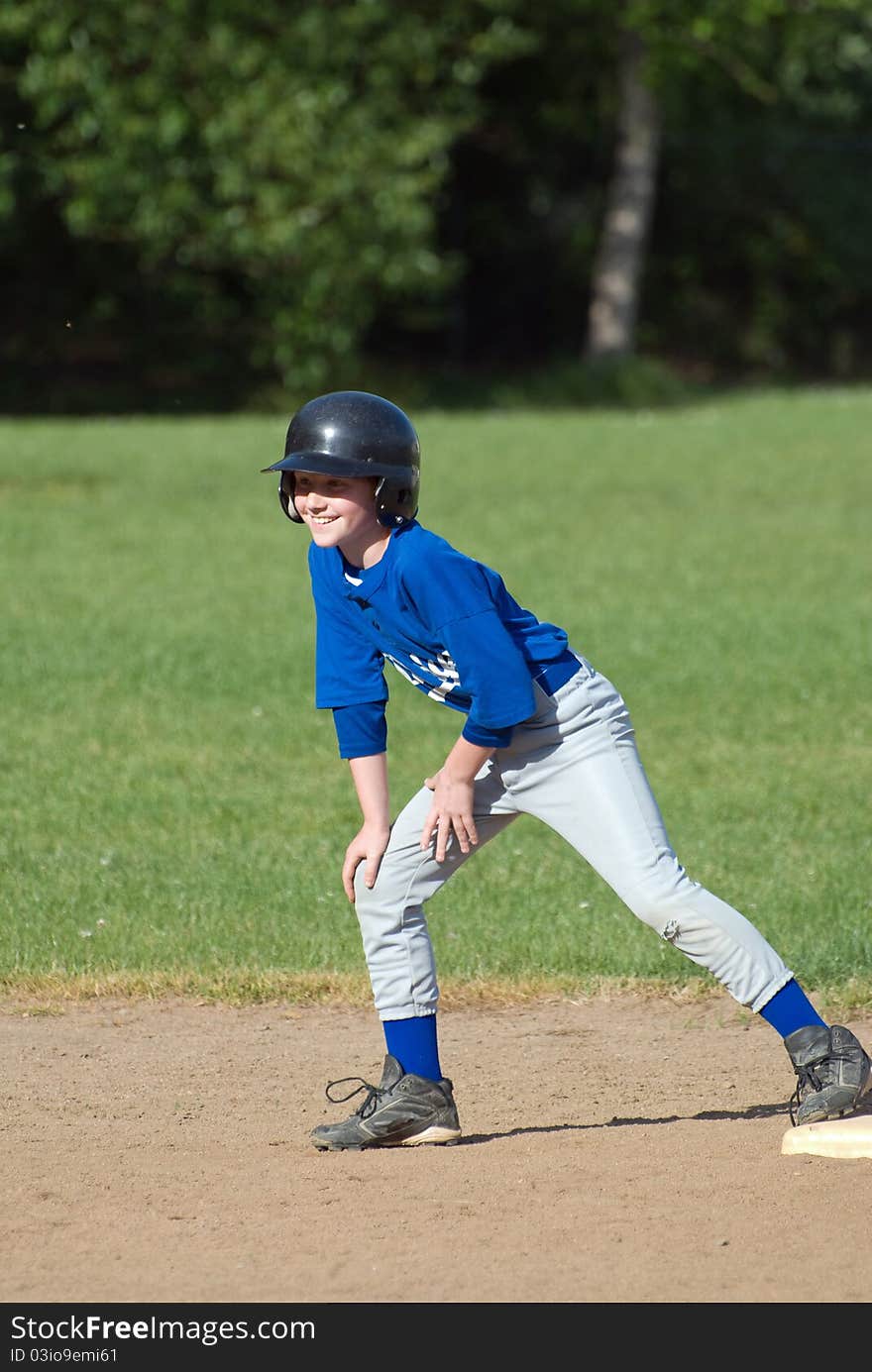 A teenage baseball player with a big smile on his facre waits for the batter to bring him home while wearing a blue and gray uniform with a black batting helmet. A teenage baseball player with a big smile on his facre waits for the batter to bring him home while wearing a blue and gray uniform with a black batting helmet