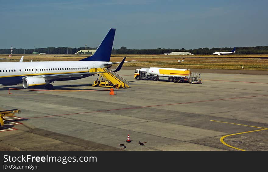 Aircraft on the ramp with a tanker truck with trailer at the rear. Ramp at rear and safety cones and ribbons around the wings. Taking off / landing aircraft on the runway in the background. Aircraft on the ramp with a tanker truck with trailer at the rear. Ramp at rear and safety cones and ribbons around the wings. Taking off / landing aircraft on the runway in the background.