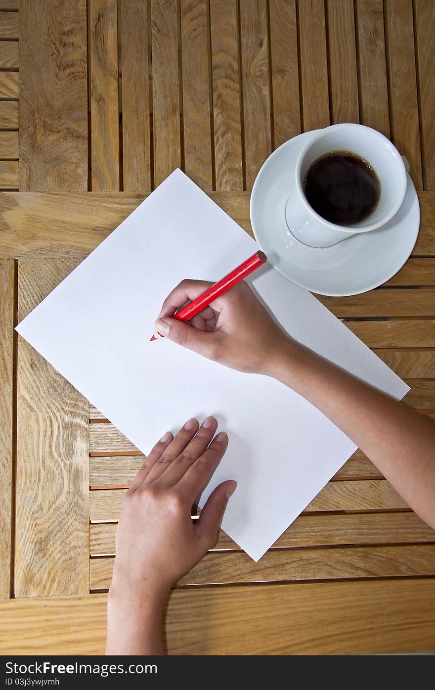 Female hand with pencil writing on a white sheet of paper. Wooden table and a cup of black coffee