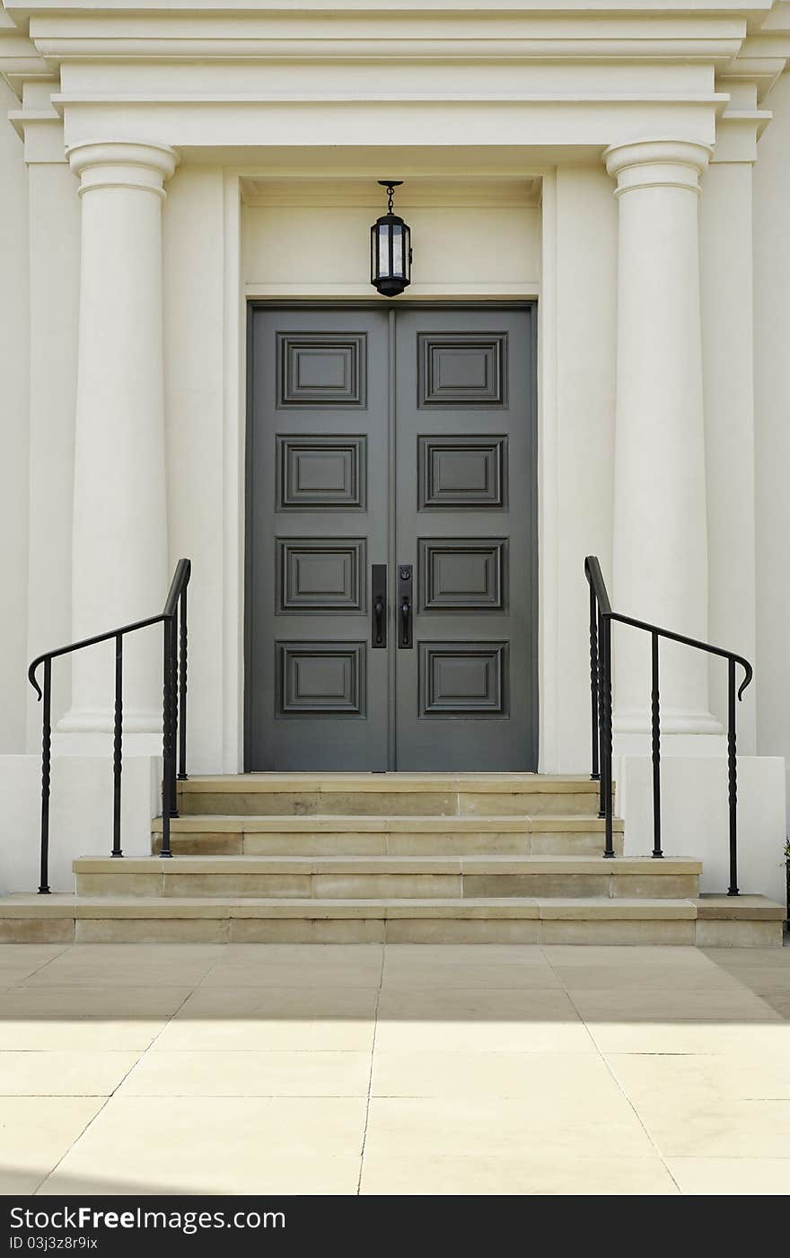 Double doors with rectangular inset patterns stand at the top of steps. There are handrails bordering the steps leading to the doors. Vertical shot.