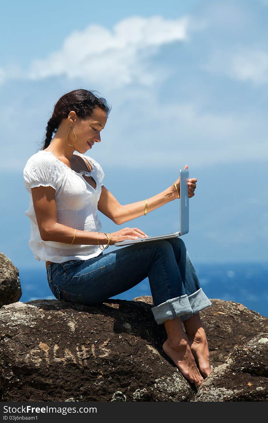 Woman works on a computer as she sits on a bluff overlooking the ocean in Maui, Hawaii. Vertical image orientation. Woman works on a computer as she sits on a bluff overlooking the ocean in Maui, Hawaii. Vertical image orientation.