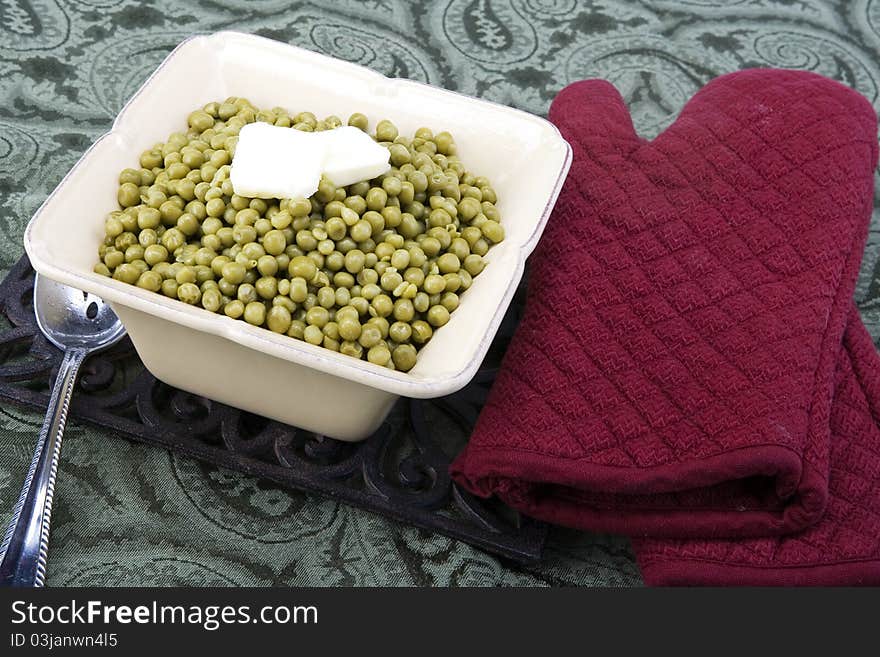 Cooked peas with butter in a ceramic dish on a green background with spoon and oven mits.