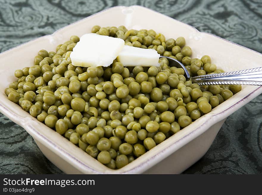 Cooked peas with butter in a ceramic dish on a green background with spoon.