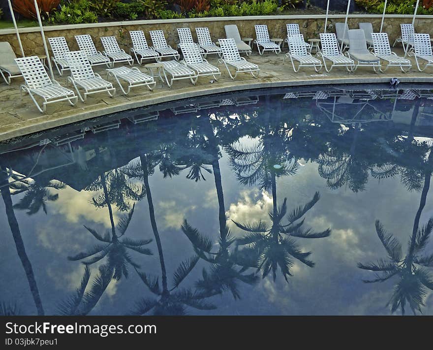 Empty chairs near a swimming pool at a Hawaiian resort, with palm trees reflected in the water. Empty chairs near a swimming pool at a Hawaiian resort, with palm trees reflected in the water.