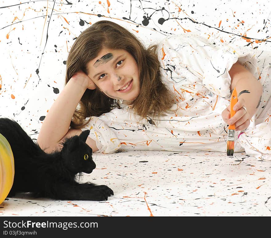 An attractive preteen painting in an orange and black paint spattered smock against a paint-spattered background. A black cat and pumpkins nearby. An attractive preteen painting in an orange and black paint spattered smock against a paint-spattered background. A black cat and pumpkins nearby.