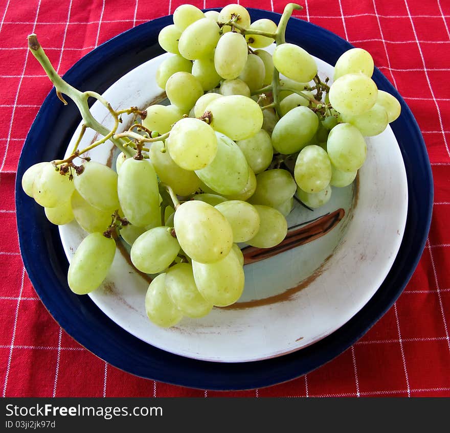 Green grapes on a white painted and a blue plate with a red and white checked tablecloth. Green grapes on a white painted and a blue plate with a red and white checked tablecloth.