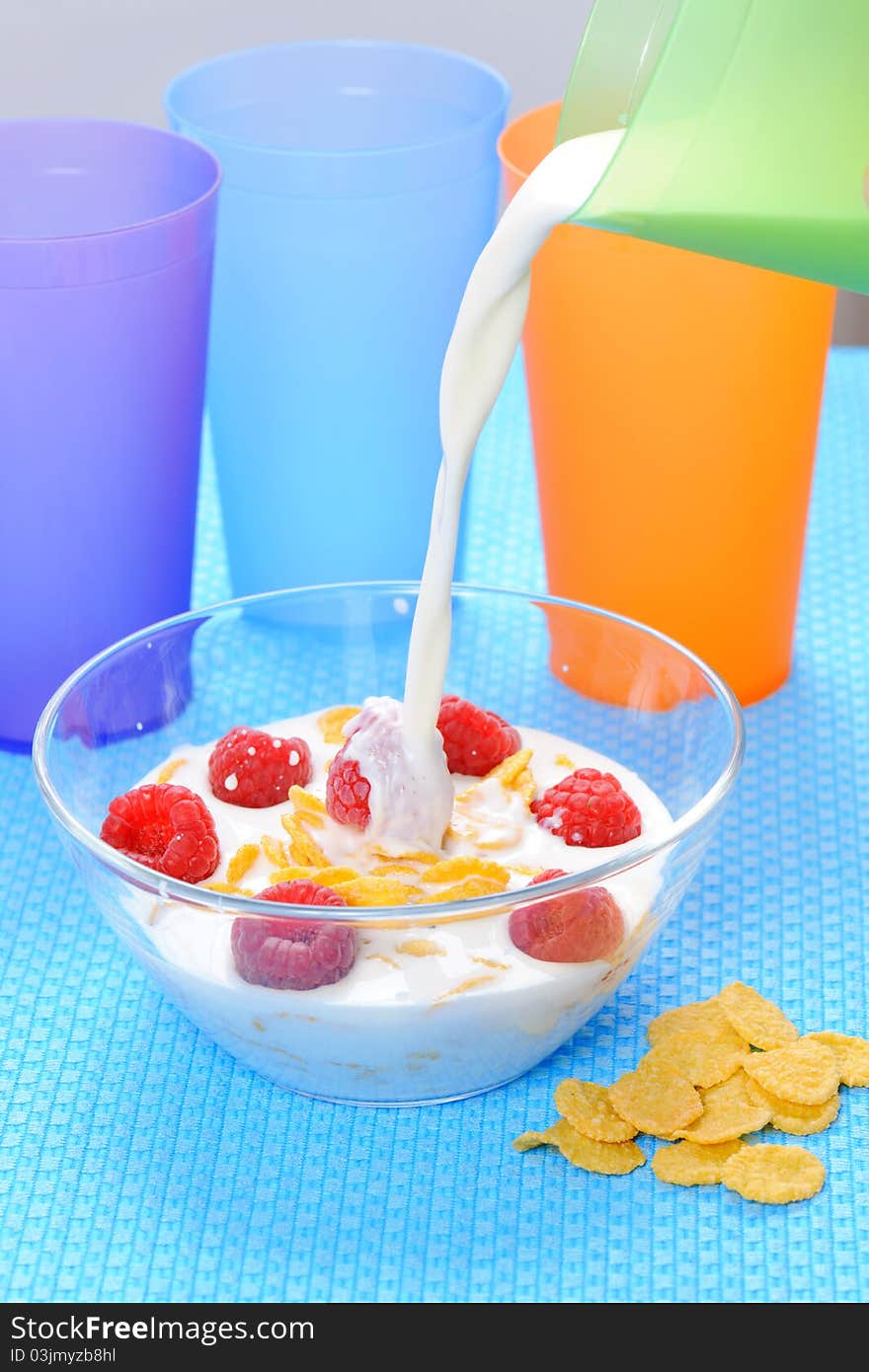 Cornflakes in glass bowl with berries, milk,  and colorful plastic cups on a blue background