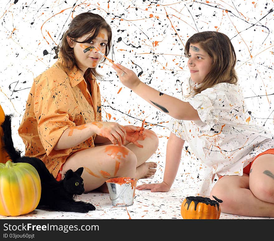 A young teen and her tween sister making a mess of themselves with black and orange paint. Pumpkins and a black cat nearby. Some motion blur on the brush painting the girl's face. A young teen and her tween sister making a mess of themselves with black and orange paint. Pumpkins and a black cat nearby. Some motion blur on the brush painting the girl's face.