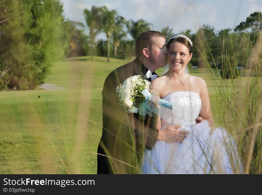 A groom kissing brides cheek outside on sunny summer day. A groom kissing brides cheek outside on sunny summer day.