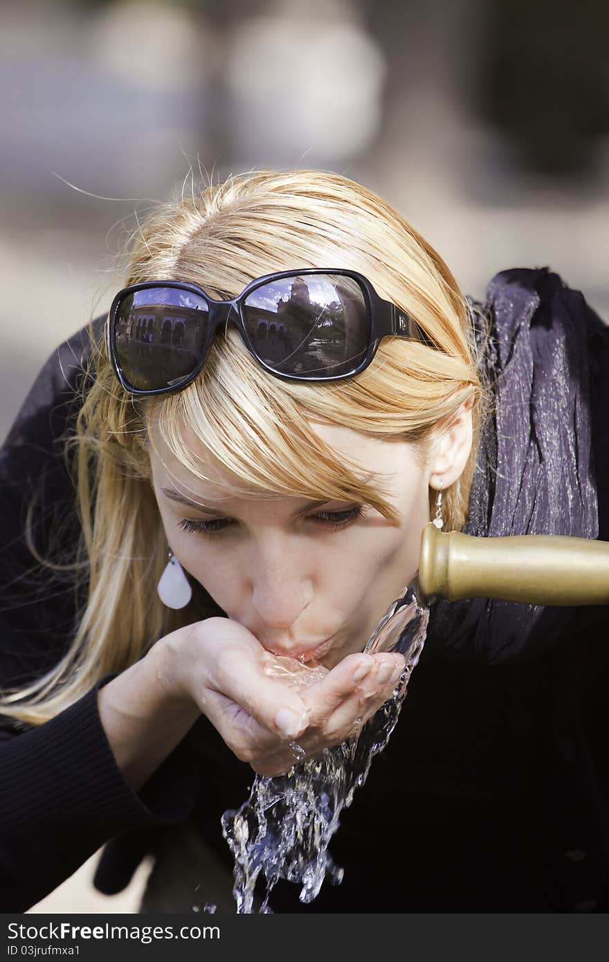 Young lady drinking tap water from the fountain in the city park on a hot summer day.