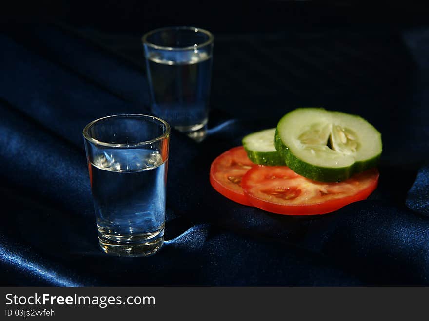 Two glasses with water or alcoholic drink and vegetables on dark blue cloth make a cold and intimate composition. Two glasses with water or alcoholic drink and vegetables on dark blue cloth make a cold and intimate composition