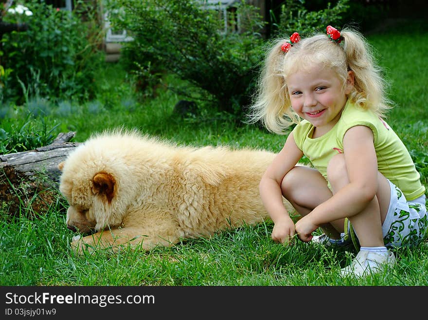 The small blonde with a naughty smile and dimples on cheeks sitting on a green lawn and nearby a dog of breed of a chow-chow