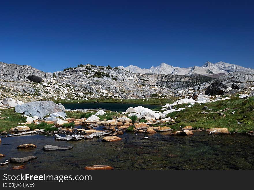 Two clear alpine lakes in the High Sierra with rugged white peaks in the background, contrasted against a dark blue sky.