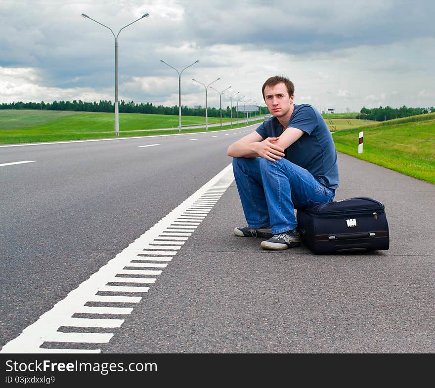 The young man sits pending on road with a suitcase