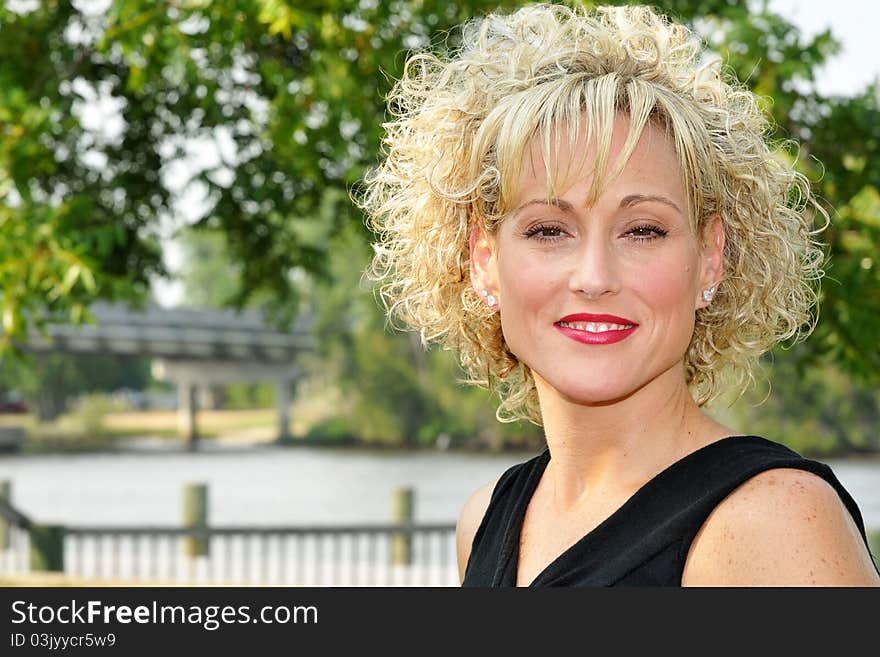 Adult blond haired woman smiling in front of a bridge, trees, and a river.