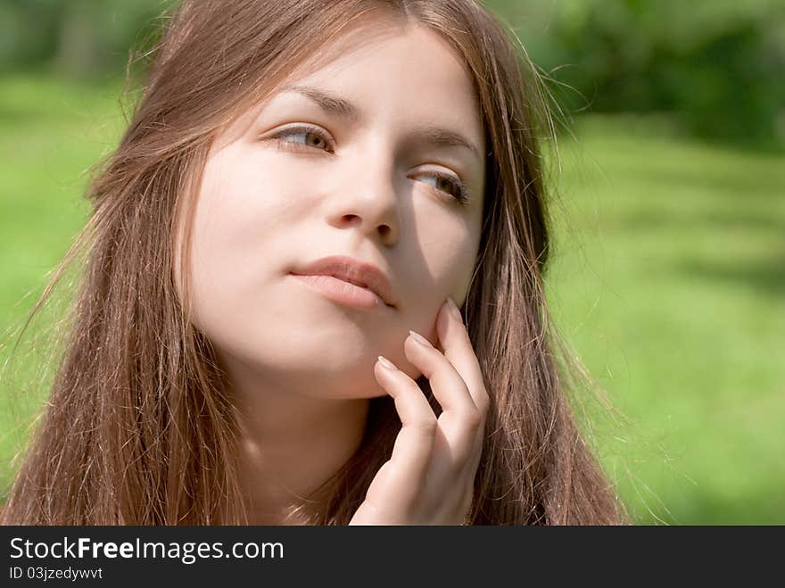 Portrait of young woman with clear skin is touching her face by hand in park on green background. Portrait of young woman with clear skin is touching her face by hand in park on green background