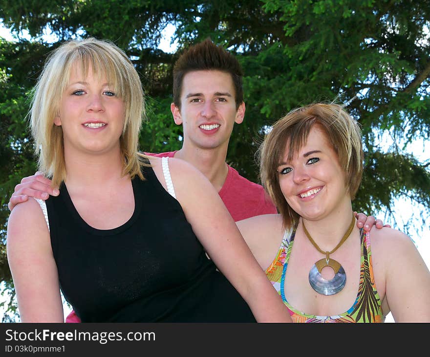 Three friends, young man with hands on shoulders of 2 young women outside with spruce tree in the backgroungd. Head and shoulders. Three friends, young man with hands on shoulders of 2 young women outside with spruce tree in the backgroungd. Head and shoulders.