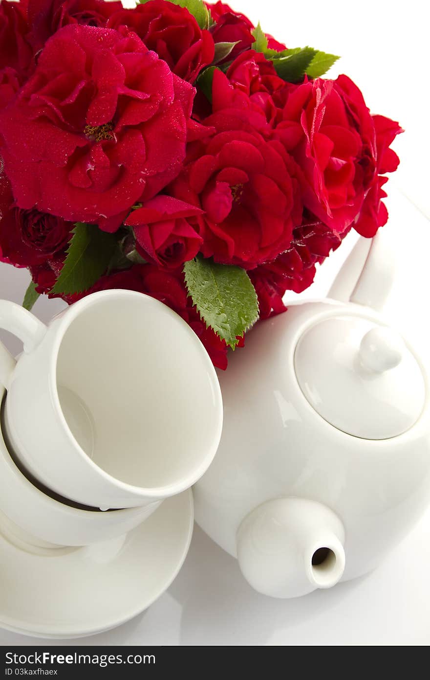 White crockery for tea kettle, two cups and saucers and a bouquet of red roses in a small iron bucket. White crockery for tea kettle, two cups and saucers and a bouquet of red roses in a small iron bucket