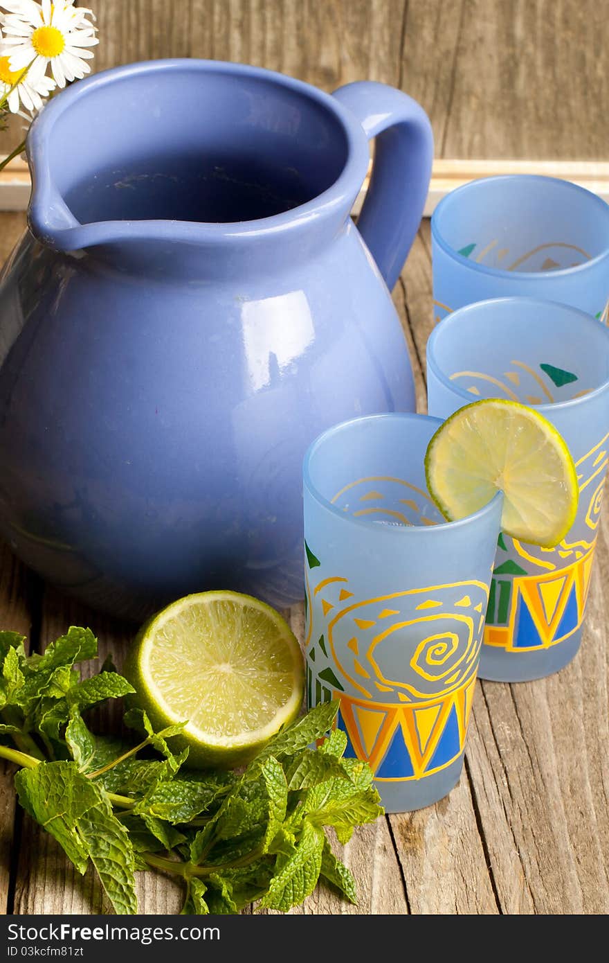 Composition with jug and three glass of lemonade, mint, half of lime and bunch of camomile on old wooden table. Composition with jug and three glass of lemonade, mint, half of lime and bunch of camomile on old wooden table