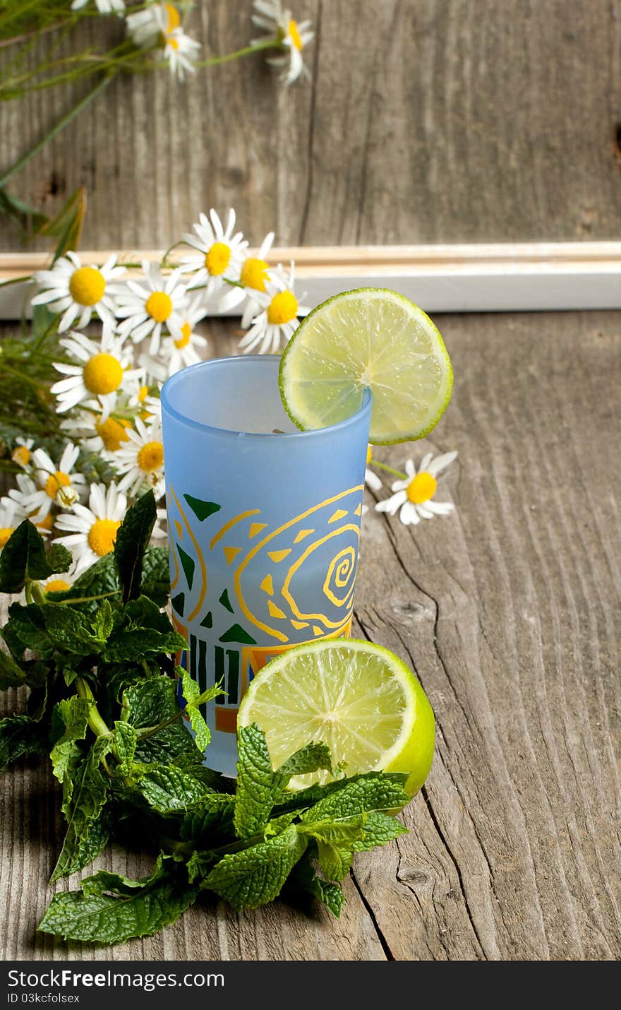 Composition with glass of lemonade, mint, half of lime and bunch of camomile on old wooden table. Composition with glass of lemonade, mint, half of lime and bunch of camomile on old wooden table