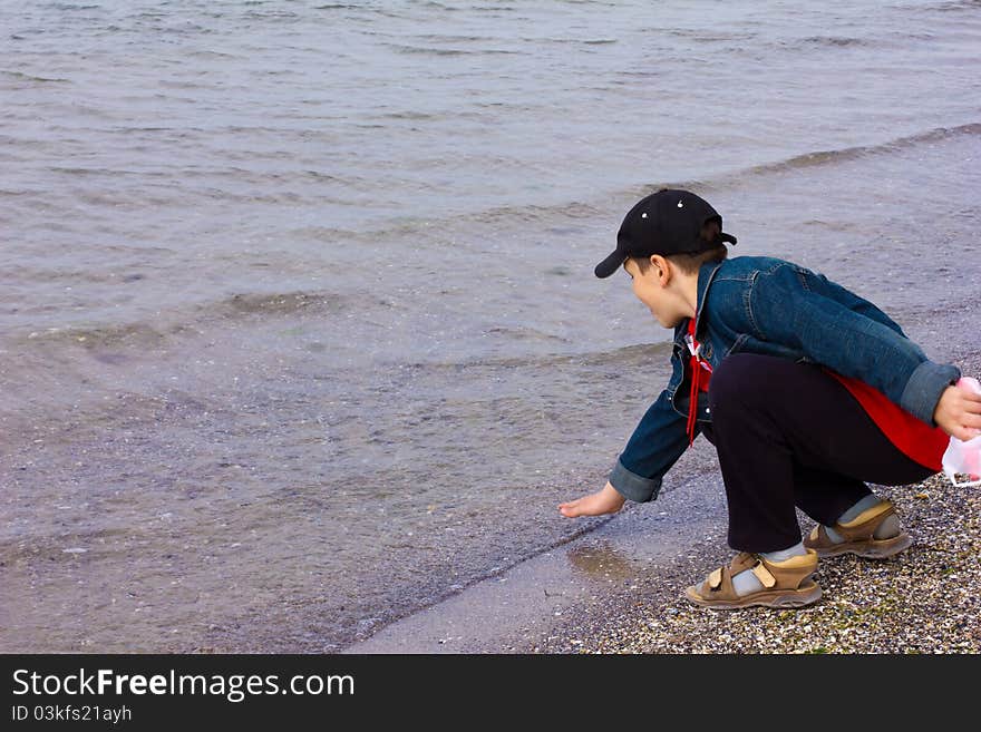 10 year old boy in denim jacket, cap, sandals squatting on the beach and the water touches his hand. 10 year old boy in denim jacket, cap, sandals squatting on the beach and the water touches his hand