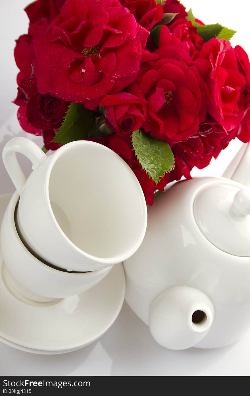 White crockery for tea kettle, two cups and saucers and a bouquet of red roses in a small iron bucket. White crockery for tea kettle, two cups and saucers and a bouquet of red roses in a small iron bucket