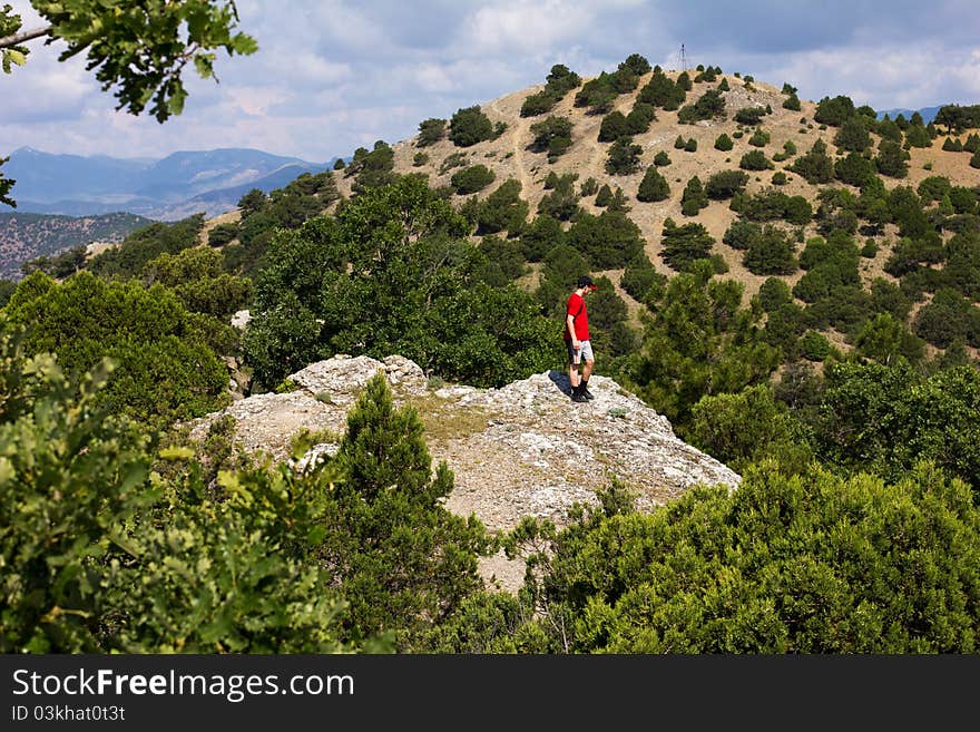 A teenager in a cap and a red shirt standing on the rocky top of a hill and looks to the next mountain. A teenager in a cap and a red shirt standing on the rocky top of a hill and looks to the next mountain