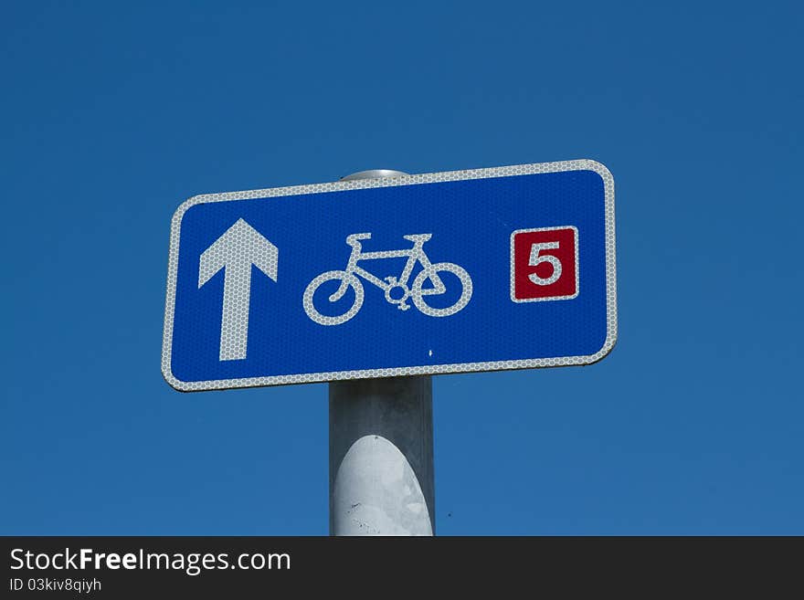 A blue sign with a white arrow and bicycle symbol and a route number five in red on a post against a blue sky. A blue sign with a white arrow and bicycle symbol and a route number five in red on a post against a blue sky.