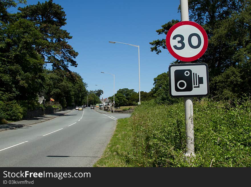 A thirty mile an hour sign and a speed camera sign on a post next to a road with cars in the distance. A thirty mile an hour sign and a speed camera sign on a post next to a road with cars in the distance.