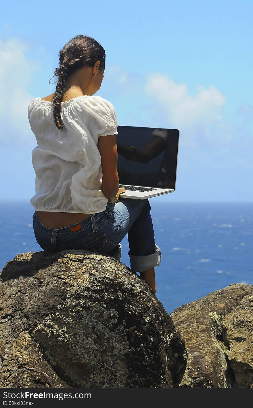 A young woman sits on a rock overlooking the ocean while working on her laptop computer. A young woman sits on a rock overlooking the ocean while working on her laptop computer