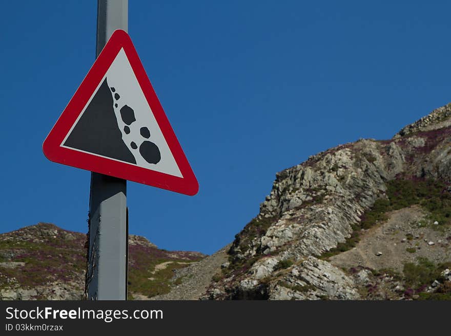 A triangular sign with red border shows falling rocks on a white background with a cliff and blue sky in the background. A triangular sign with red border shows falling rocks on a white background with a cliff and blue sky in the background.