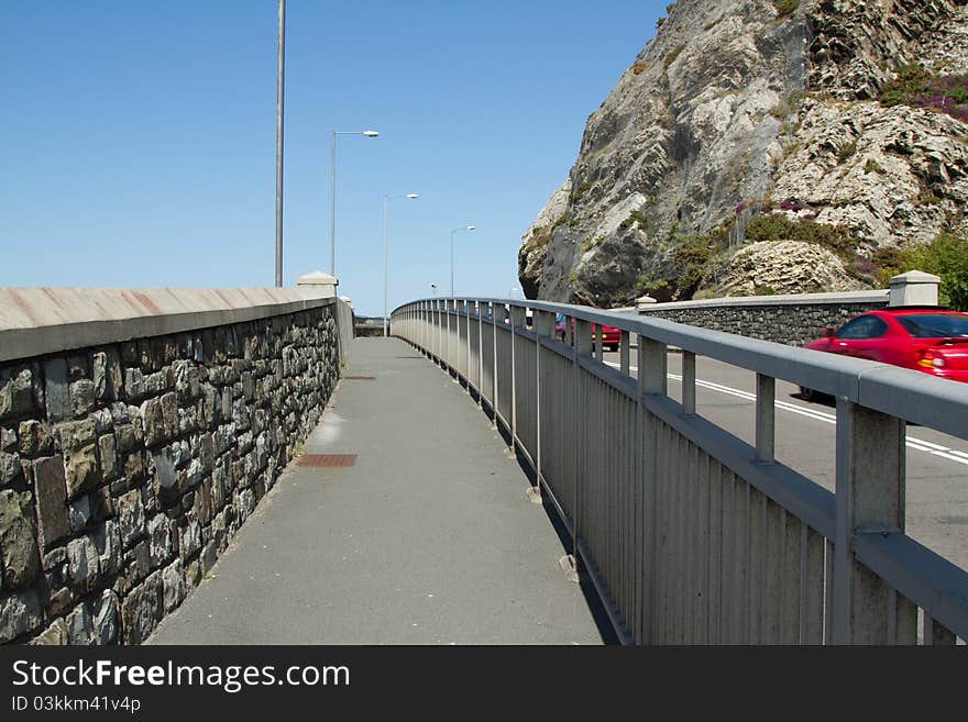 A pavement with a stone wall and lamp posts and metal railings separating the road with traffic. A pavement with a stone wall and lamp posts and metal railings separating the road with traffic.