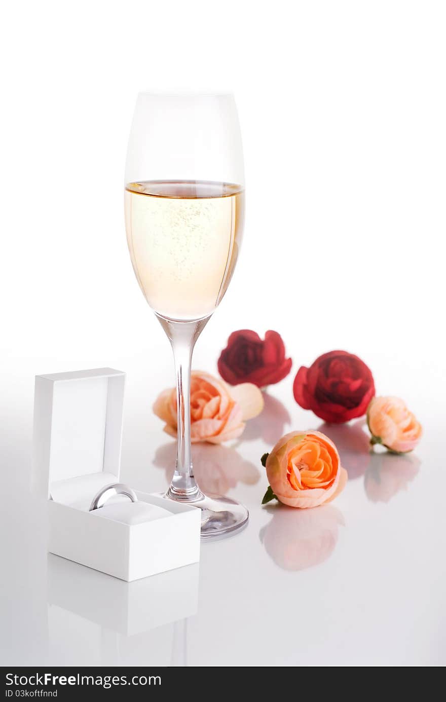 Champagne glass filled with Champagne surrounded by pink and red roses and a white jewelry box with a ring in it on a white background. Champagne glass filled with Champagne surrounded by pink and red roses and a white jewelry box with a ring in it on a white background