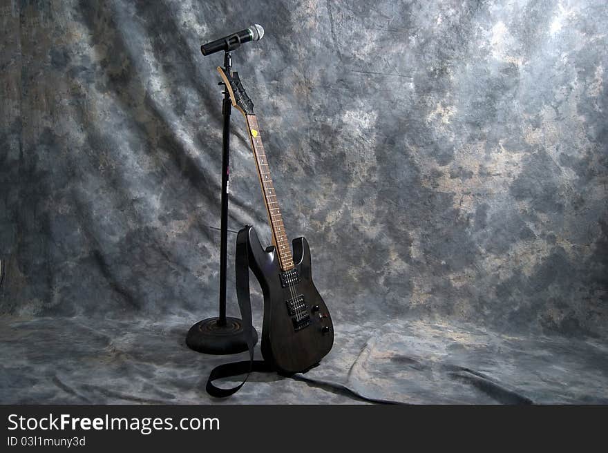 A black electric guitar is resting on a microphone stand with a microphone attached. A black electric guitar is resting on a microphone stand with a microphone attached.