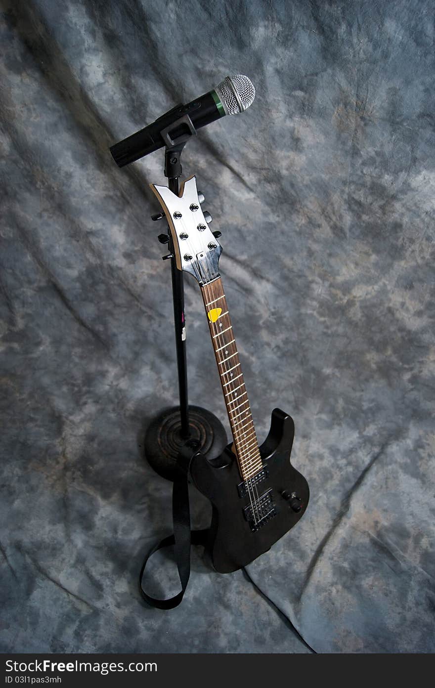 A black electric guitar is resting on a microphone stand with a microphone attached. A black electric guitar is resting on a microphone stand with a microphone attached.