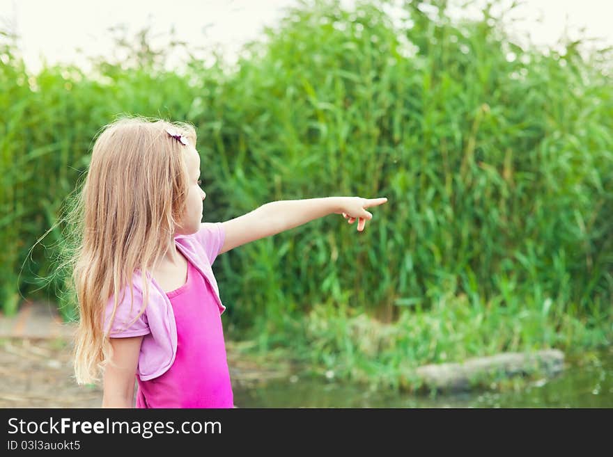 Blonde little girl shows forefingers aside near river with canes in summer