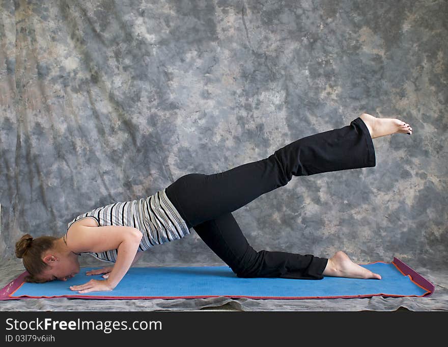 Young woman on yoga mat in Yoga posture sunbird pose with left leg lifted, against a grey background in profile, facing left lit by diffused sunlight. Young woman on yoga mat in Yoga posture sunbird pose with left leg lifted, against a grey background in profile, facing left lit by diffused sunlight.