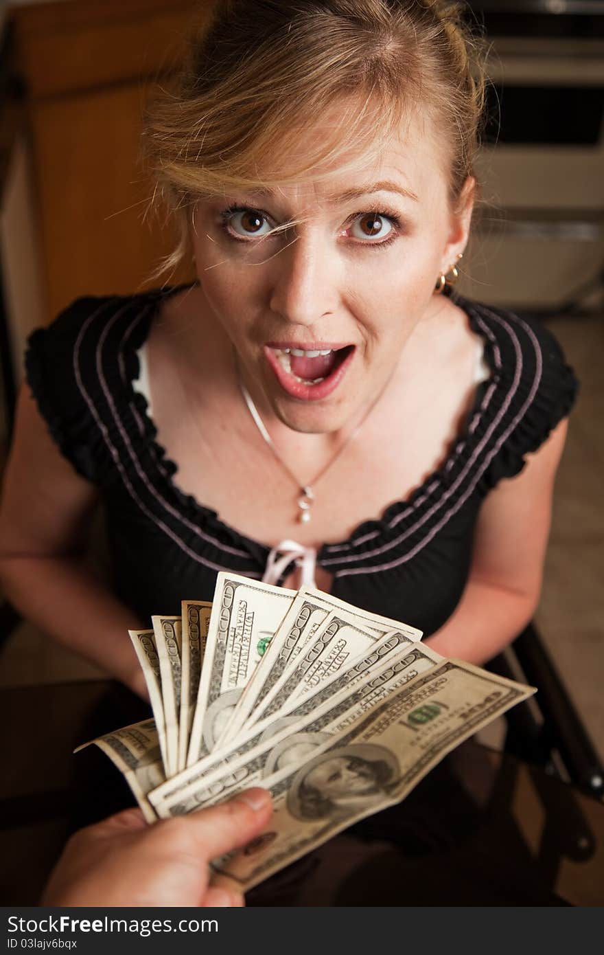 Blond woman in casual attire with a shocked expression sitting at home with hand giving lots of 100 dollar bills. Blond woman in casual attire with a shocked expression sitting at home with hand giving lots of 100 dollar bills