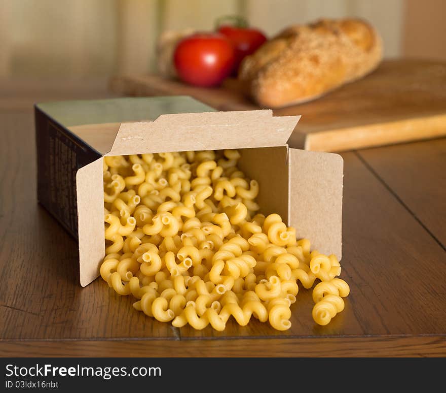 Uncooked corkscrew shaped pasta spilling from the box, with a loaf of Italian bread and tomatoes in the background. Uncooked corkscrew shaped pasta spilling from the box, with a loaf of Italian bread and tomatoes in the background