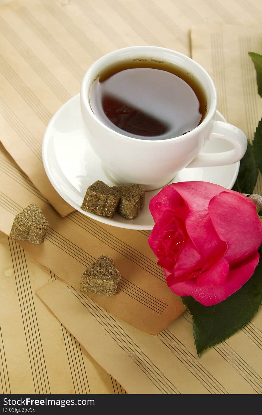 White cup of tea next to sugar and is a beautiful red rose lying on sheets of old paper sheet music. White cup of tea next to sugar and is a beautiful red rose lying on sheets of old paper sheet music