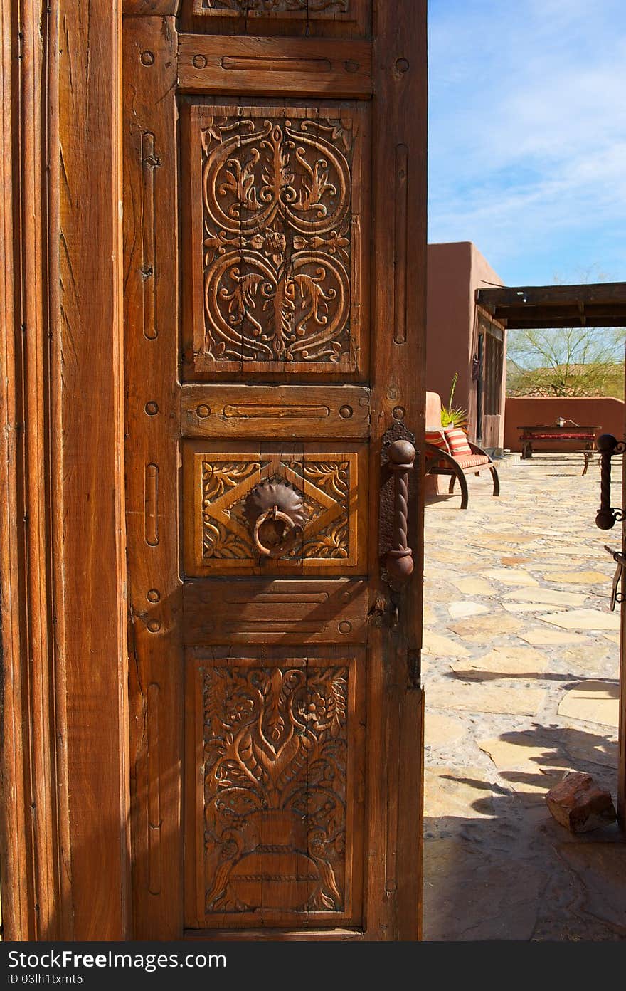 A wooden, ornate, opened door with metal knocker leads to a inner courtyard with stone and seating. A wooden, ornate, opened door with metal knocker leads to a inner courtyard with stone and seating