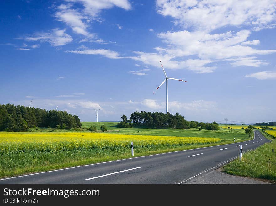 Two wind turbines standing in the green field near the emty road.