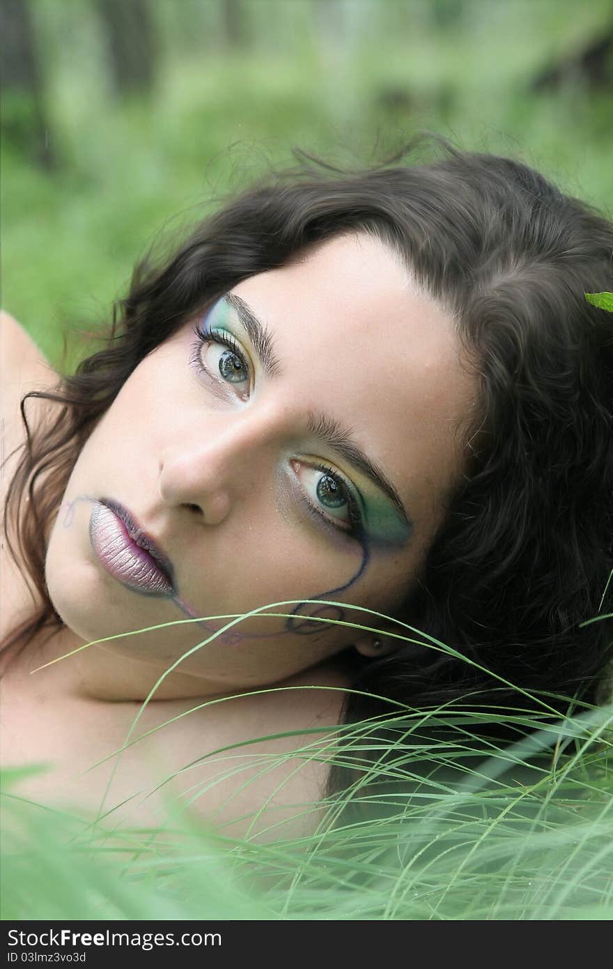 Young woman laying in the grass after the rain, a pale rainbow of makeup on her face.