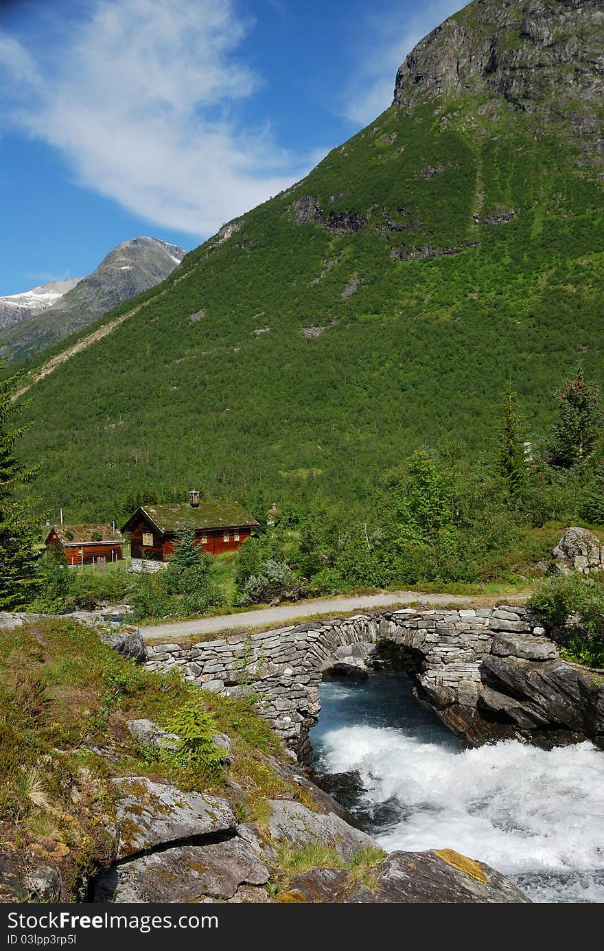 Small wooden houses with greenroof are located under the green mountain. Rapid river is running under stone bridge in the foreground. Small wooden houses with greenroof are located under the green mountain. Rapid river is running under stone bridge in the foreground.