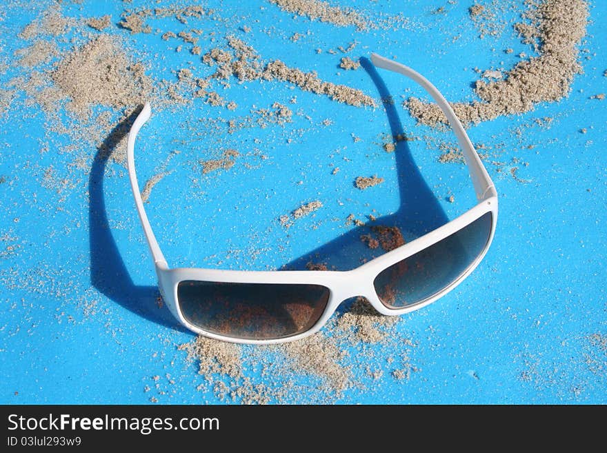 White sunglasses sitting on a blue backround with sand.