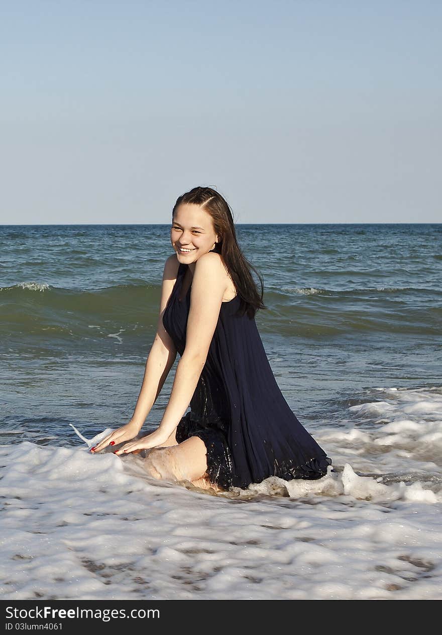 Beautiful young girl in black wet dress sits in water of the sea