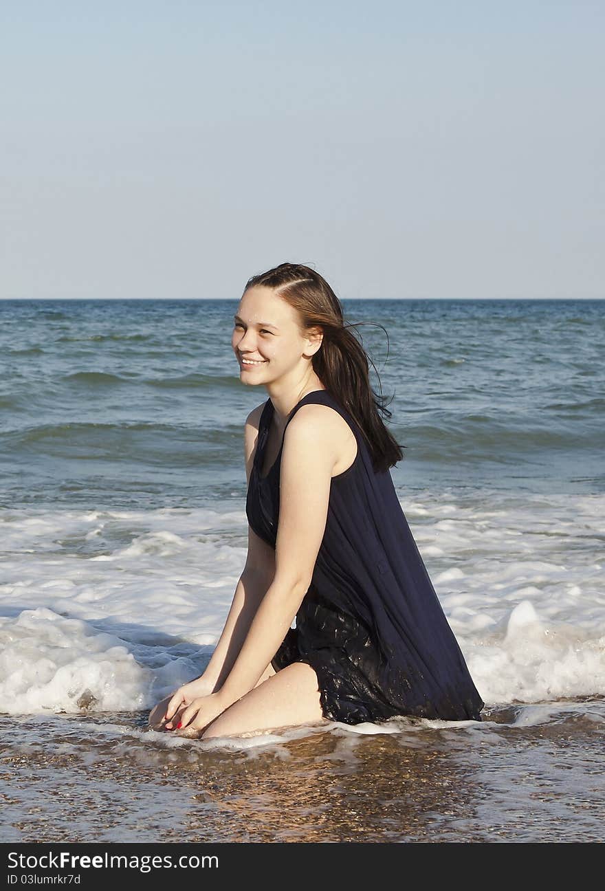 Beautiful young girl in black wet dress sits in water of the sea