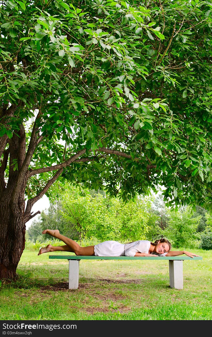 Pretty barefooted woman with is laying on a bench under the green tree. She is slightly smiling and looking at the camera. Pretty barefooted woman with is laying on a bench under the green tree. She is slightly smiling and looking at the camera