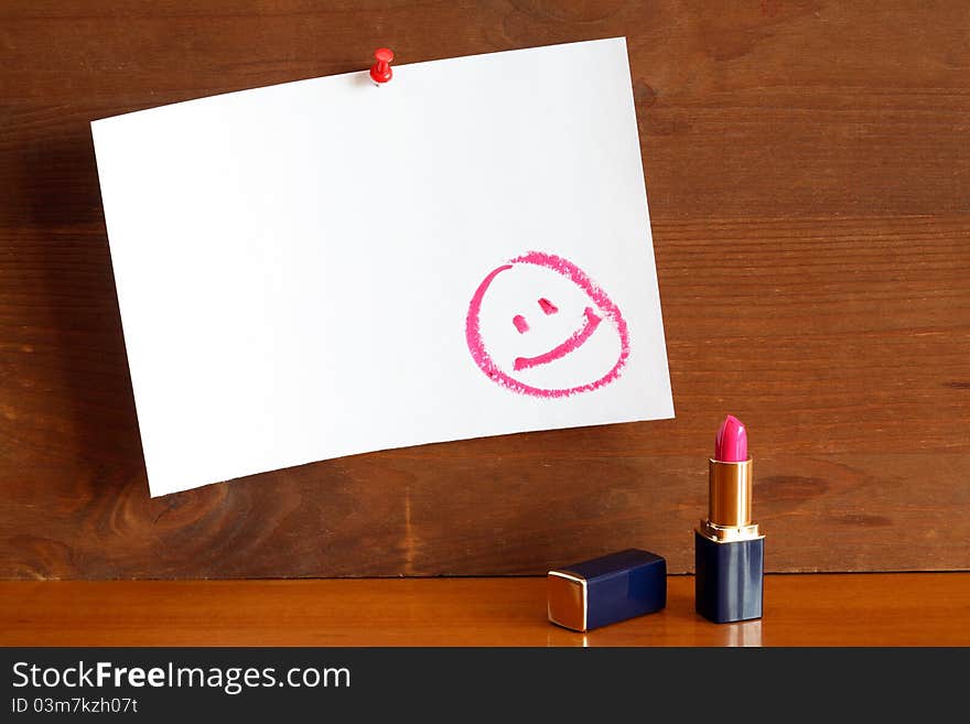 Lipstick standing near blank paper sheet with drawn smiling face. Paper is hanging on wooden wall. Lipstick standing near blank paper sheet with drawn smiling face. Paper is hanging on wooden wall