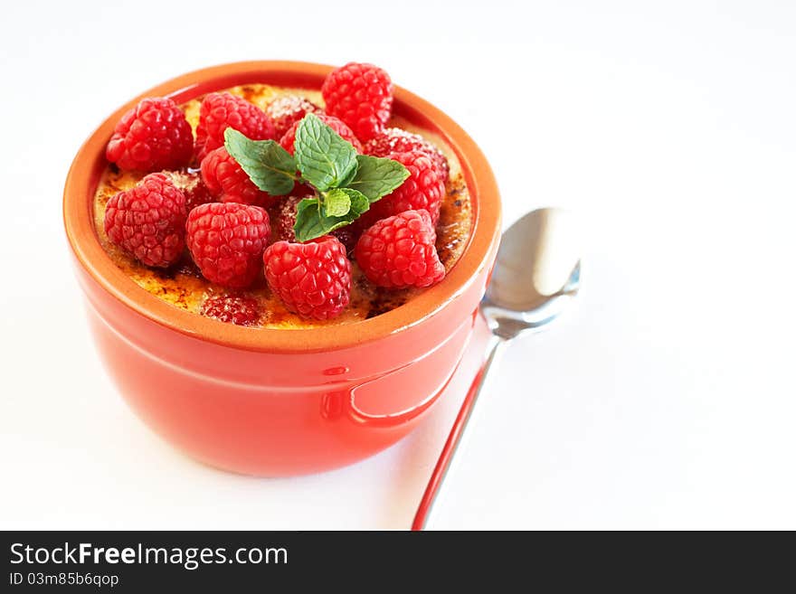 French creme brulee dessert with raspberries and mint covered with caramelized sugar in red terracotta ramekin on white background